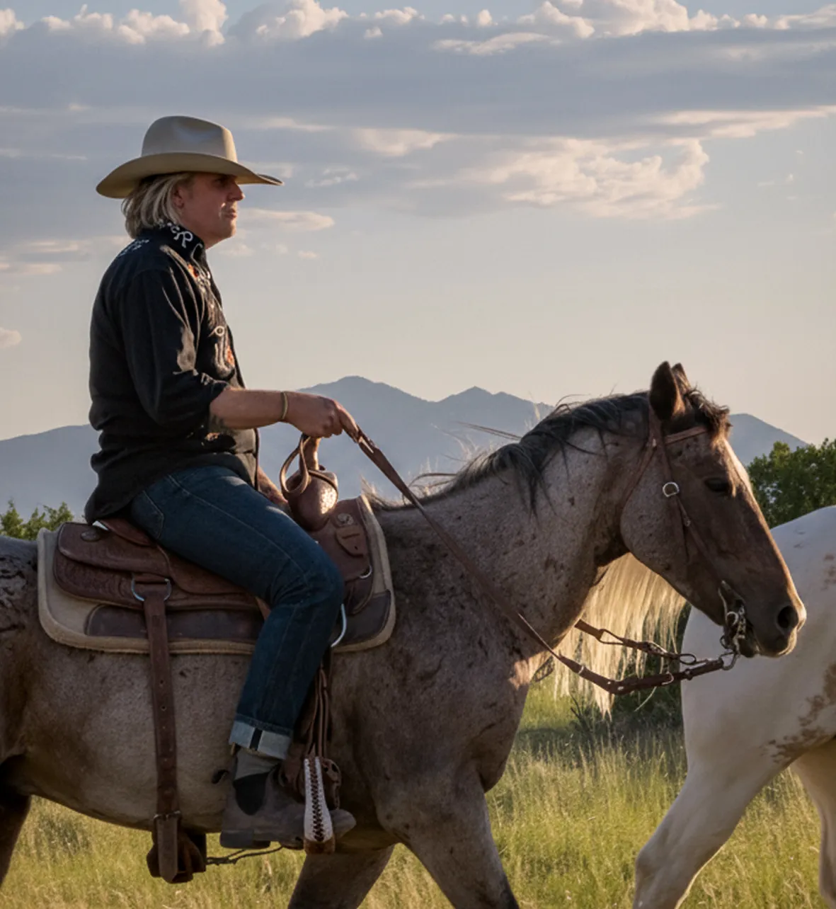 A cowboy rides a horse through a grassy landscape with mountains in the background during a scenic sunset.