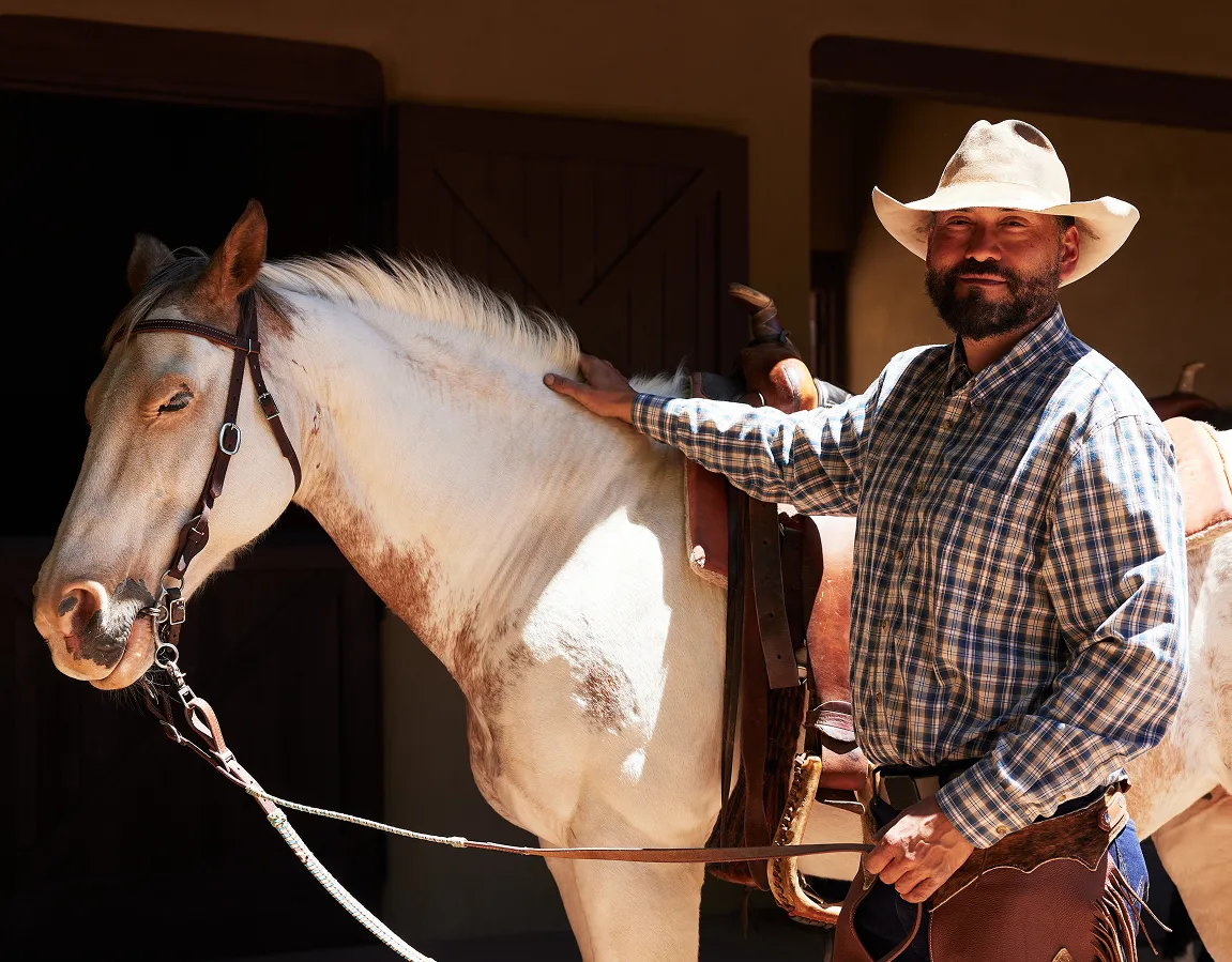 A cowboy in a plaid shirt stands next to a white horse, ready for a ride in a stable environment.