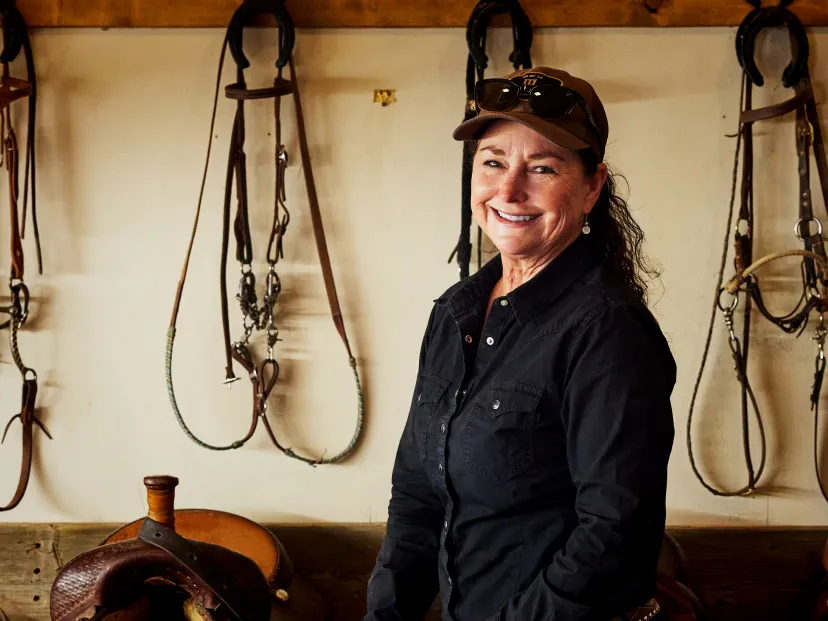 Smiling woman in a black shirt stands in front of horse tack, showcasing a warm and welcoming equestrian environment.