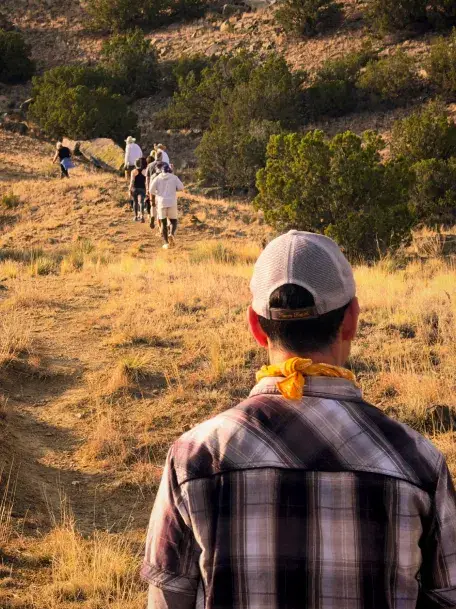 A group of hikers follows a dirt path through grassland and trees, with one person in a plaid shirt and cap in the foreground.