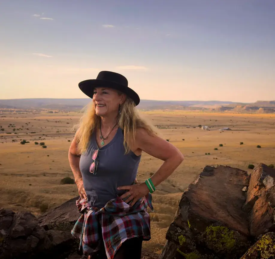 A smiling woman in a hat stands on rocks overlooking a vast landscape under a clear sky, exuding a sense of adventure.