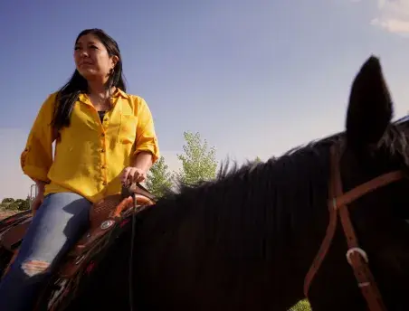 A woman in a yellow shirt confidently rides a horse under a clear blue sky.