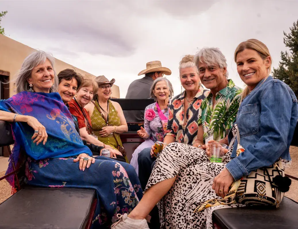 A group of smiling friends enjoying a casual gathering outdoors, dressed in colorful attire and sharing drinks.