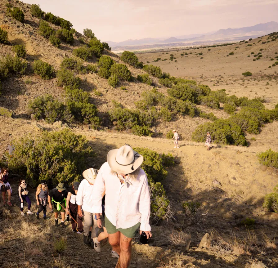 A group of hikers ascends a rocky trail surrounded by shrubs and mountains under a clear sky.