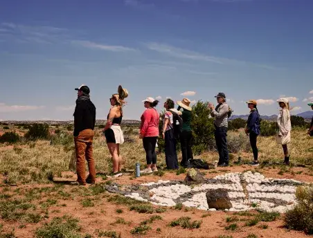 A group of people stands in a line outdoors, observing a circular stone formation under a clear blue sky.