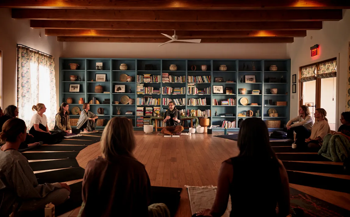 A serene meditation session with participants seated in a circle, focused on a speaker in a cozy, book-filled room.
