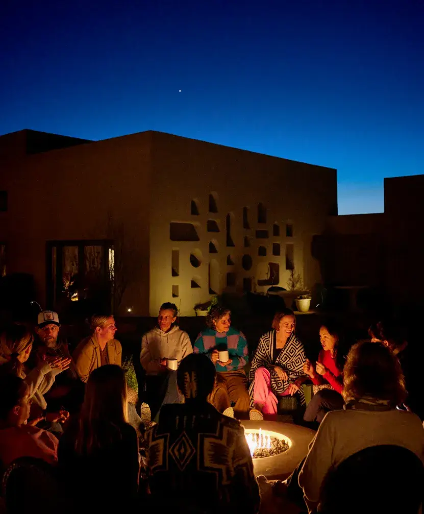 A group of people gathered around a fire pit, enjoying conversation under a twilight sky.