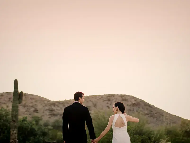 A couple in formal attire holds hands, walking towards a scenic desert backdrop at sunset.