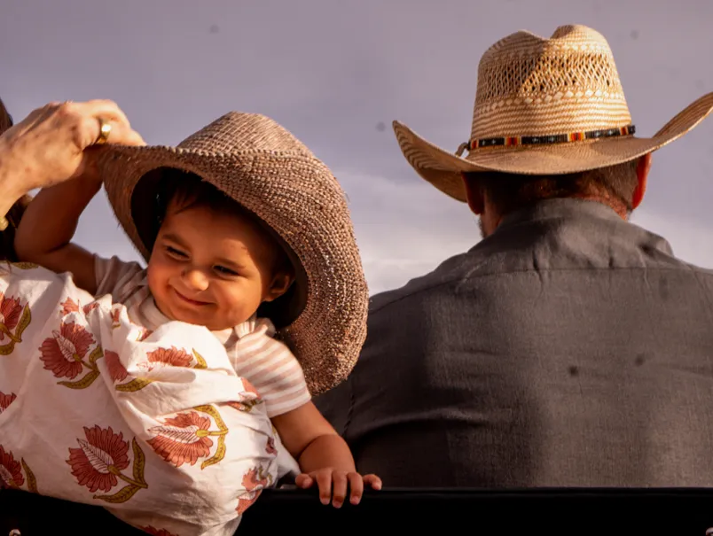A joyful child wearing a straw hat is playfully reaching for the hat of an adult behind them, both enjoying a sunny moment.