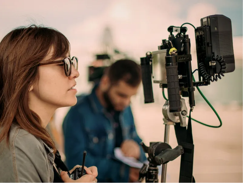 A woman with sunglasses operates filming equipment while a man takes notes in the background.