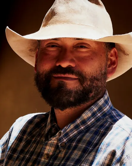 A smiling man with a beard wears a cowboy hat and plaid shirt, set against a warm, softly lit background.