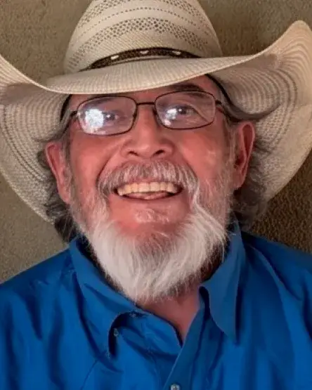 Smiling older man in a cowboy hat and glasses, wearing a blue shirt, against a neutral background.
