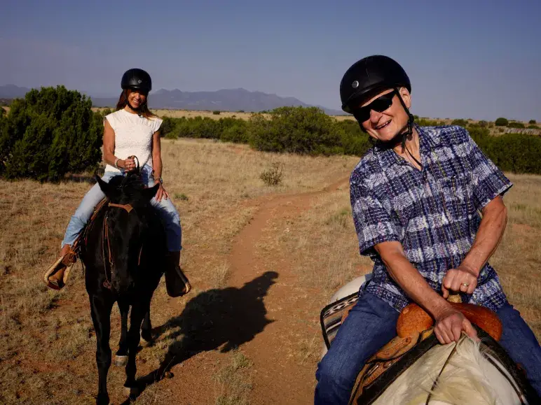 Two riders on horseback enjoy a scenic trail in a sunny outdoor setting, wearing helmets and smiling.