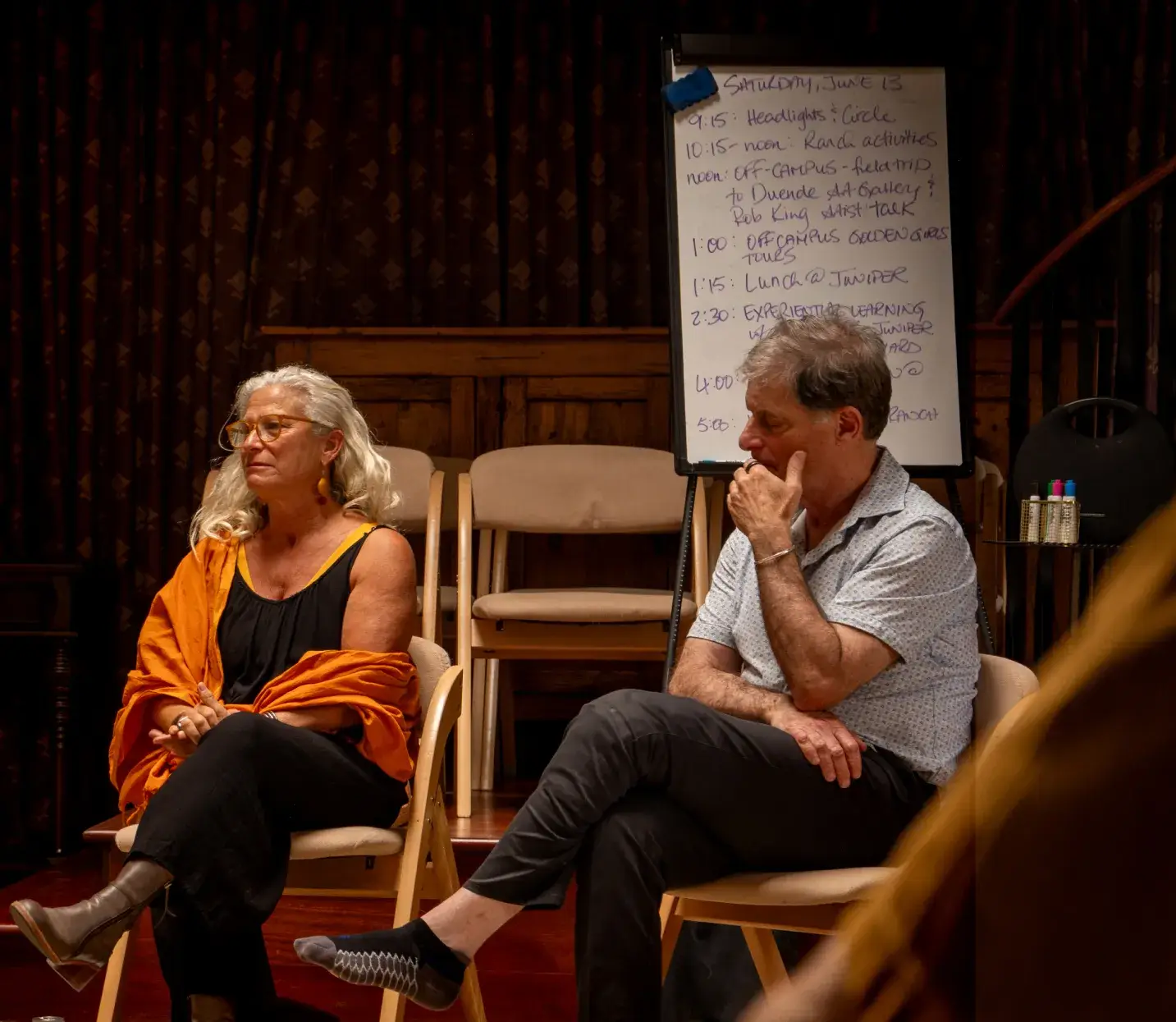 Two participants engaged in a discussion during a workshop, with a schedule on a whiteboard in the background.