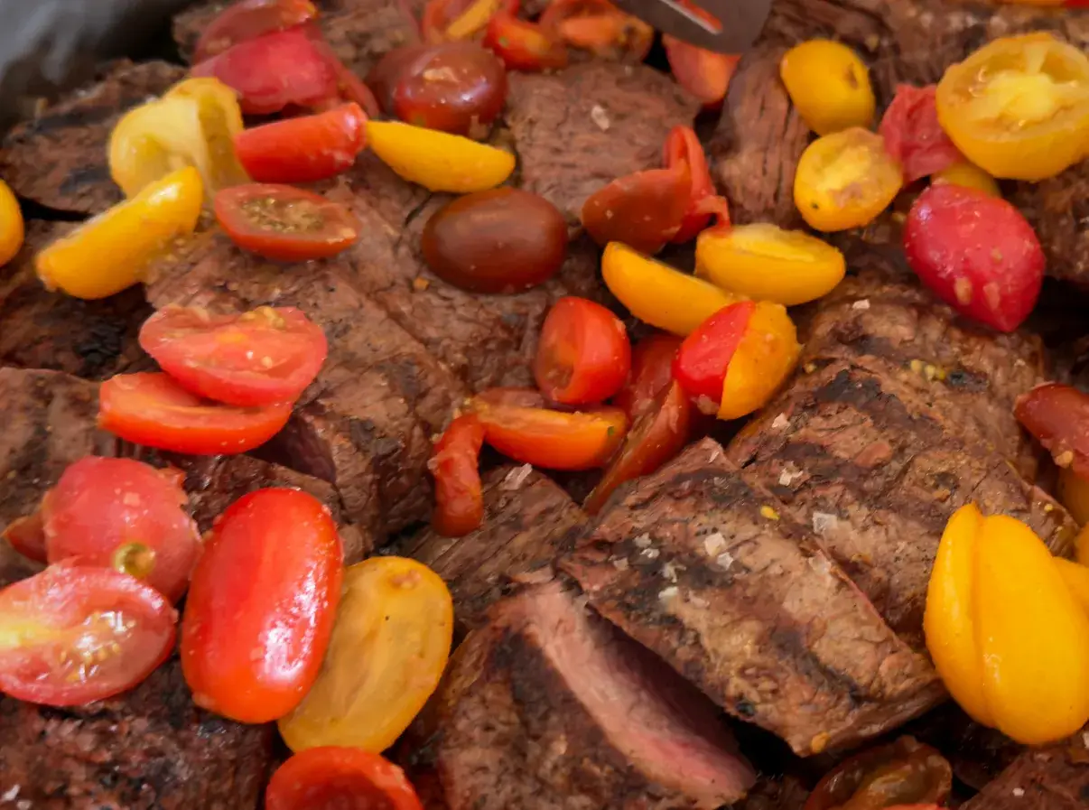 Sliced grilled beef topped with colorful cherry tomatoes, showcasing a flavorful dish ready for serving.