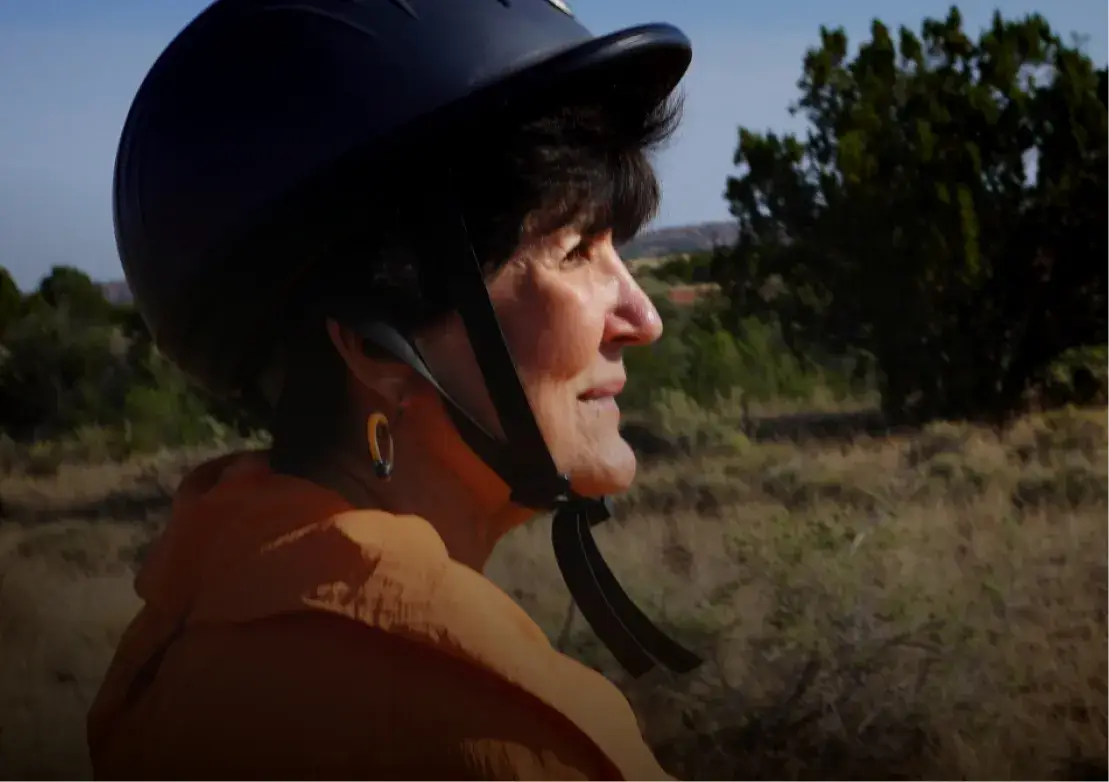 A woman wearing a riding helmet gazes thoughtfully into the distance amidst a natural landscape.