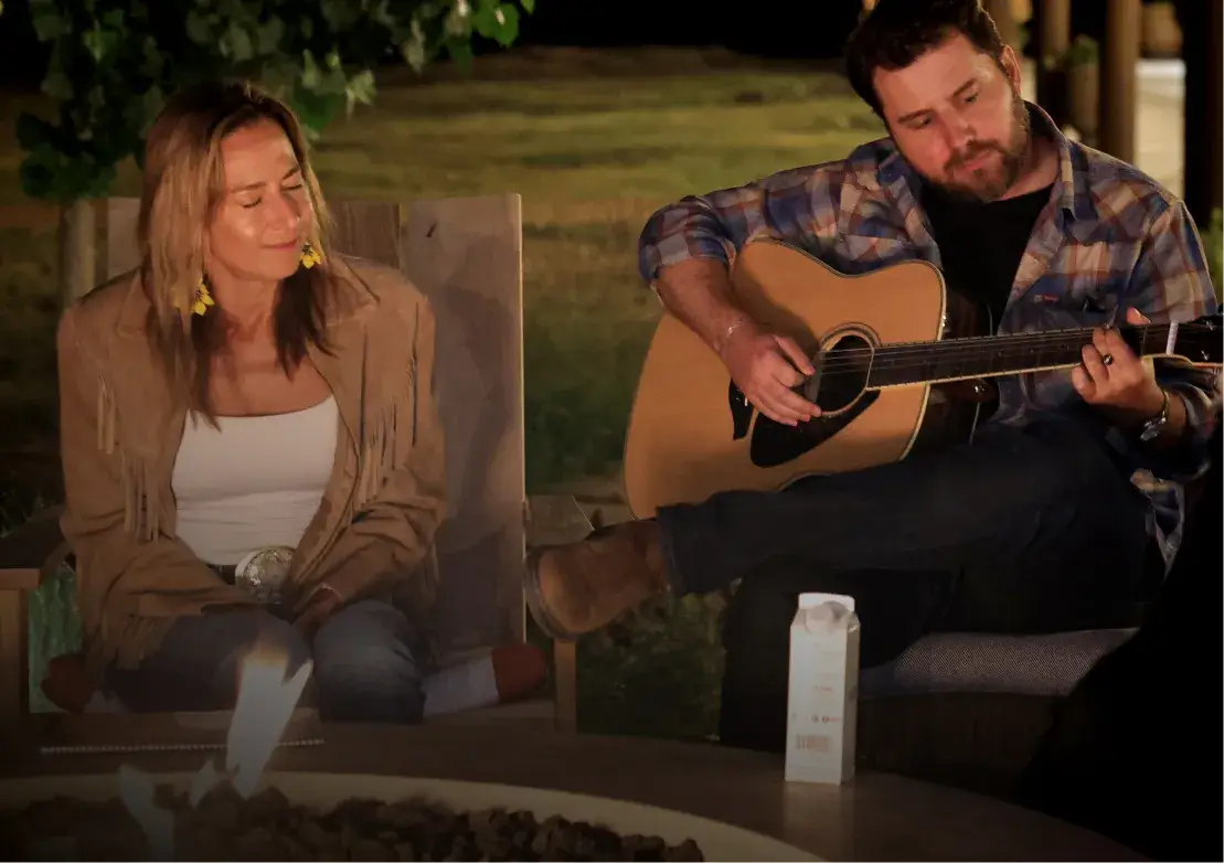A woman listens intently as a man plays guitar by a campfire, creating a cozy, intimate outdoor atmosphere.