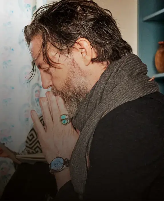 A man with a beard and long hair, wearing a scarf, meditates with hands clasped in prayer against a softly lit background.