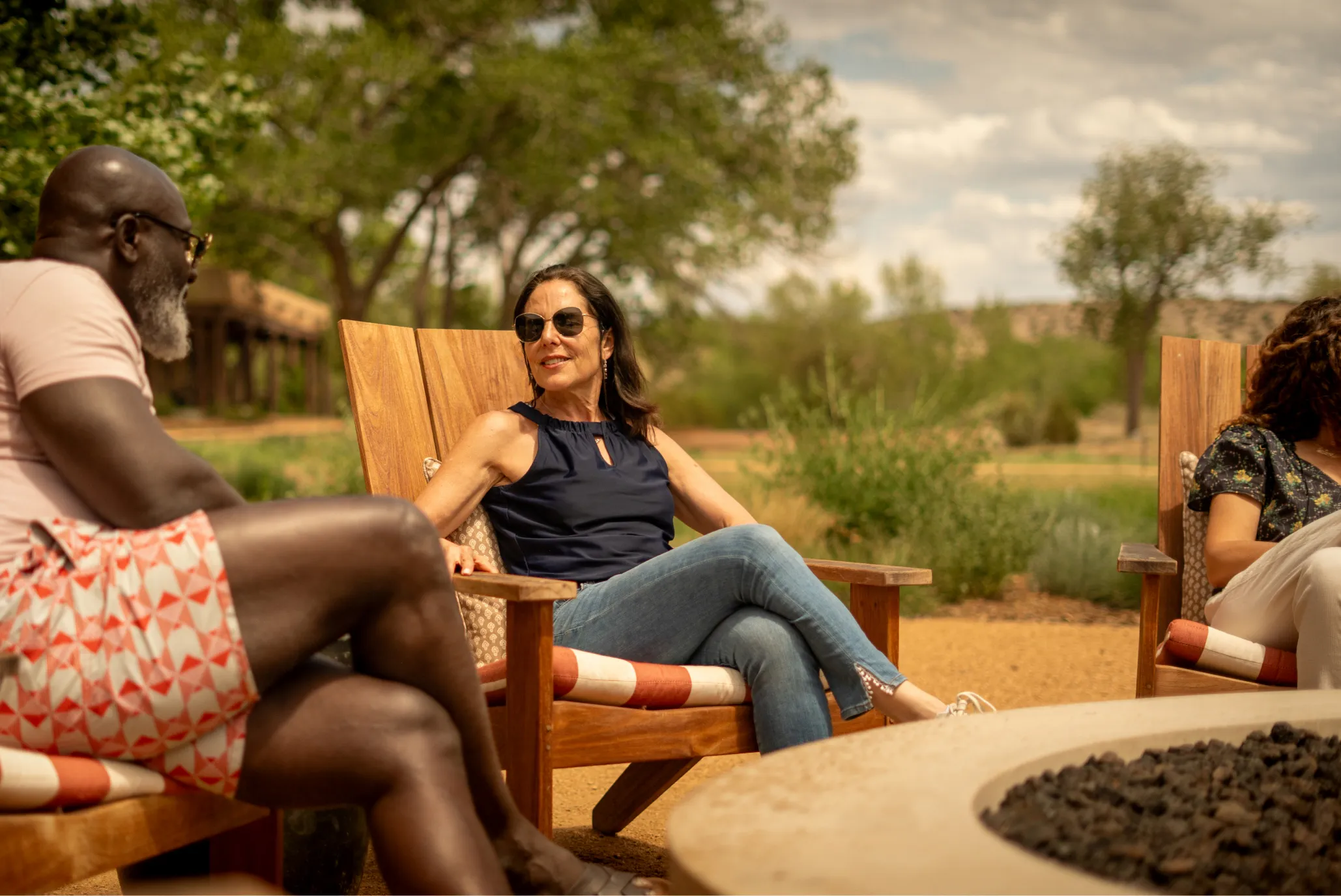 Three friends enjoy a relaxed conversation outdoors in comfortable chairs near a fire pit, surrounded by greenery.