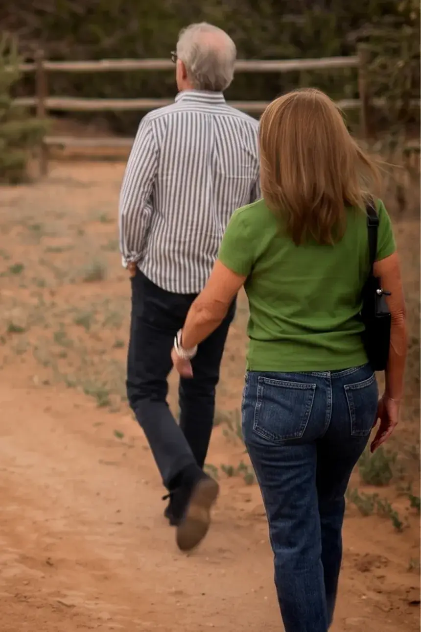 A couple walks along a dirt path, surrounded by greenery and a wooden fence, enjoying a leisurely stroll together.
