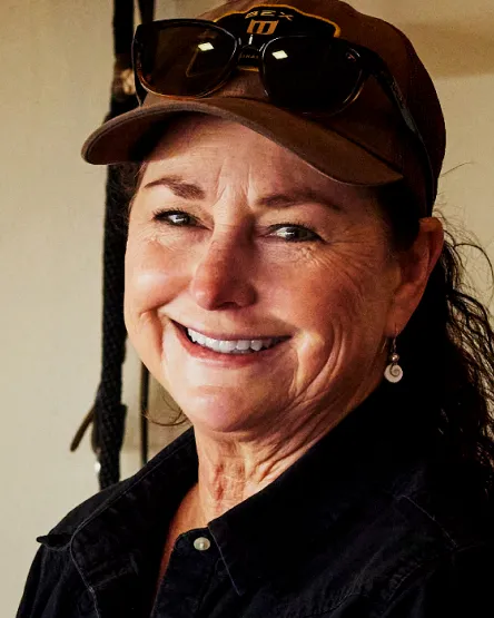 Smiling woman wearing a cap and sunglasses, exuding warmth and friendliness in a rustic setting.