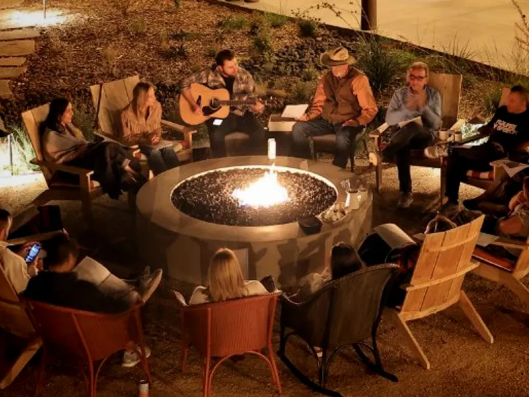 A group of people gather around a fire pit at night, enjoying music and conversation in a cozy outdoor setting.
