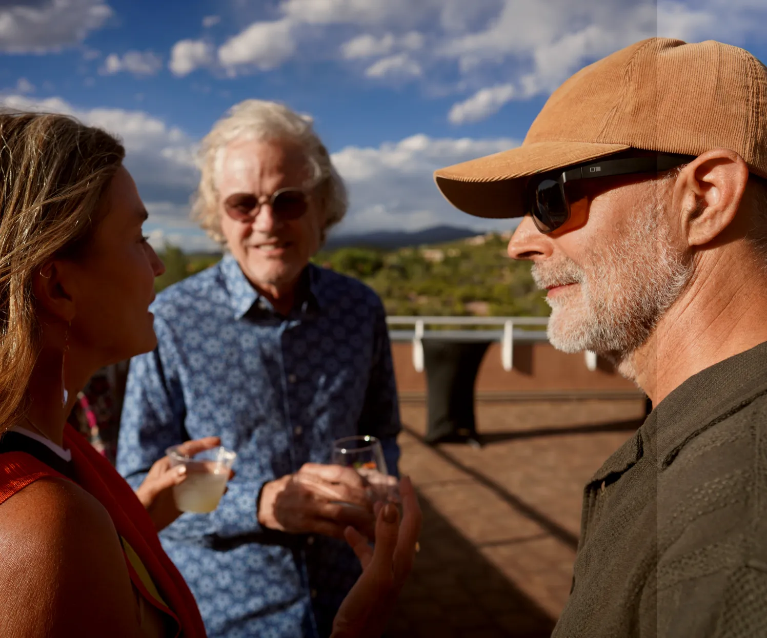 Three people engaged in conversation outdoors, enjoying drinks with a scenic background of clouds and greenery.