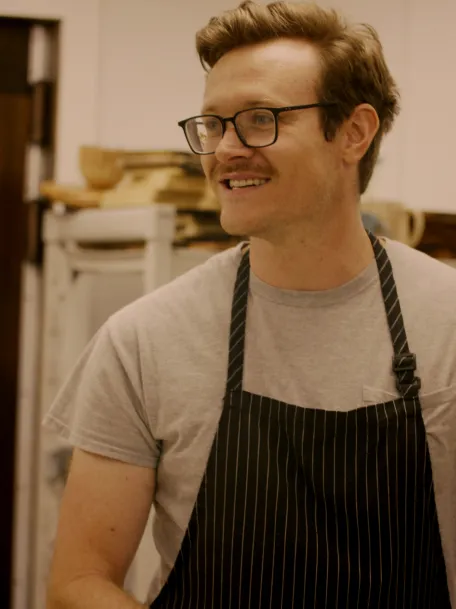 A smiling chef in a striped apron stands in a kitchen, engaging with others while preparing food.