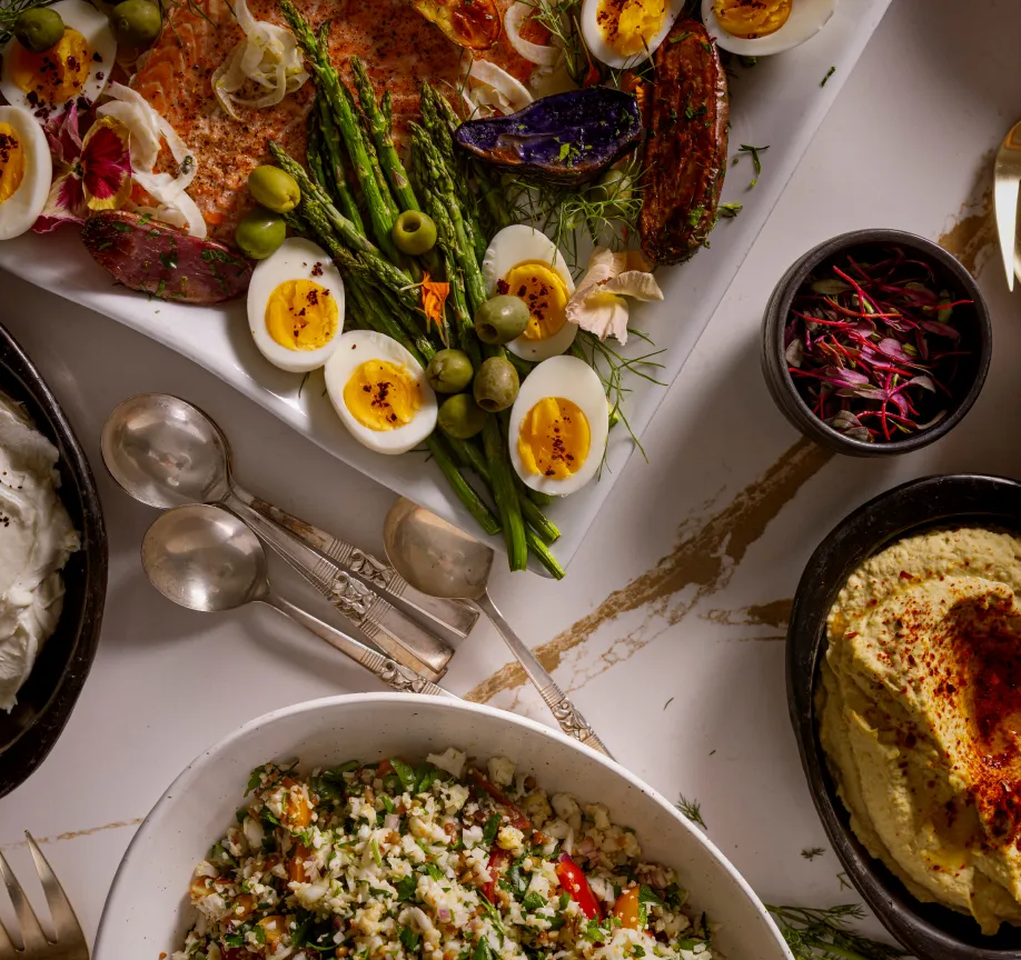 A vibrant spread of salmon, asparagus, deviled eggs, and sides like hummus and grain salad, garnished with herbs.