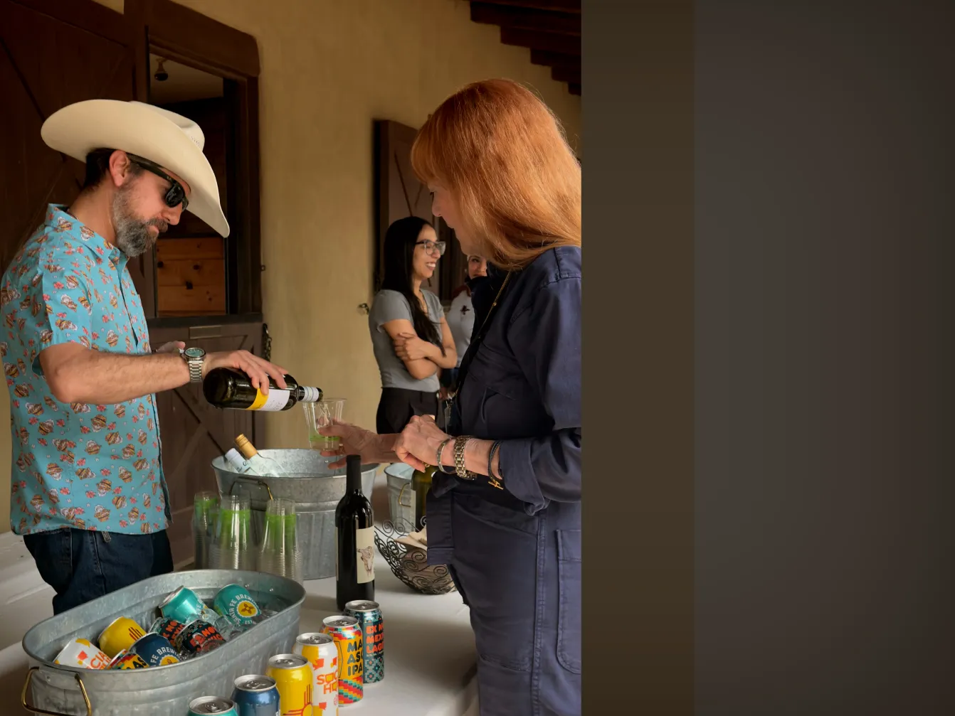 A man in a cowboy hat pours wine for a woman at a casual gathering with various drinks on display.