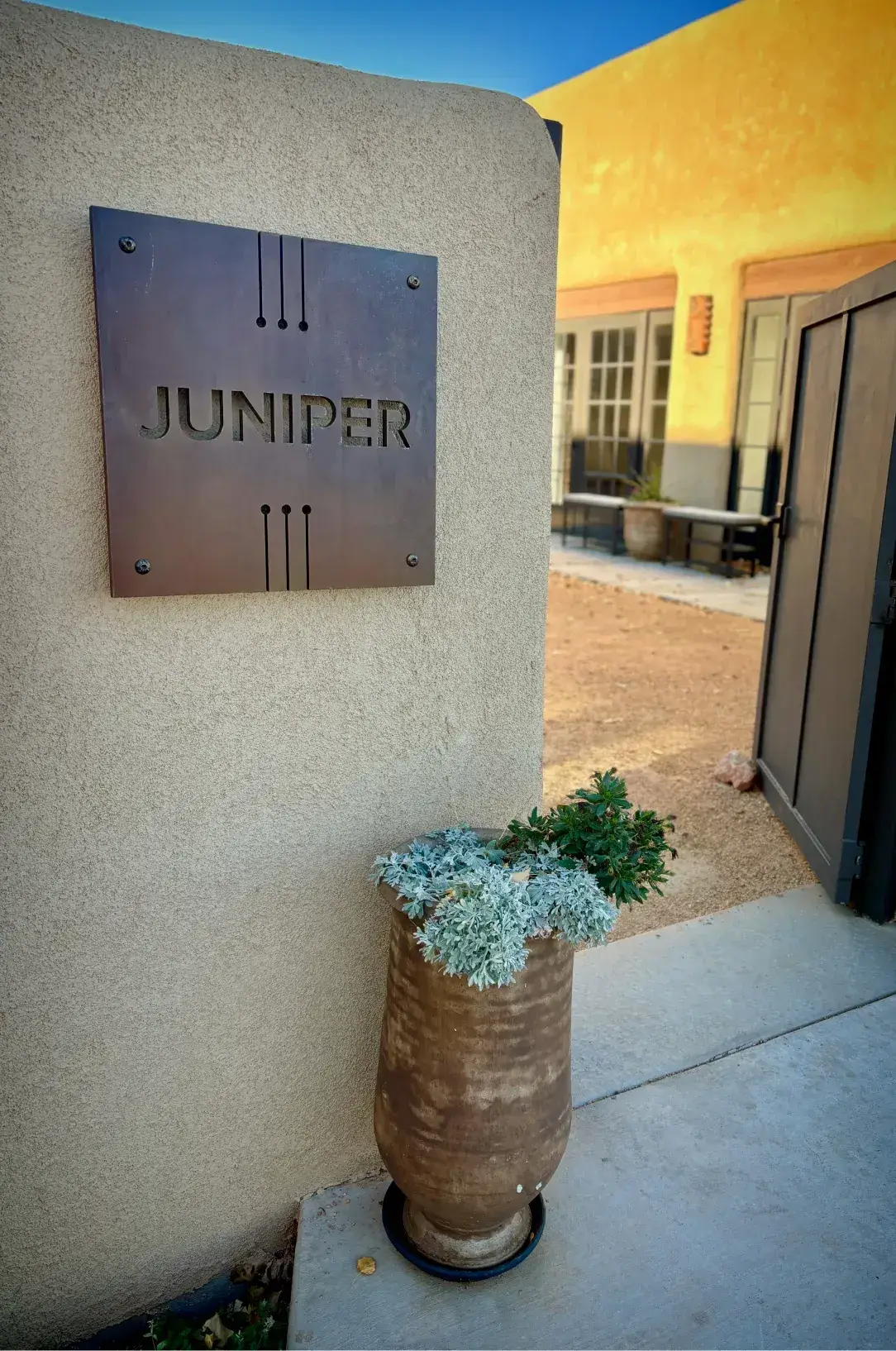 Juniper sign mounted on a wall beside a decorative planter with green plants, highlighting the entrance to a building.