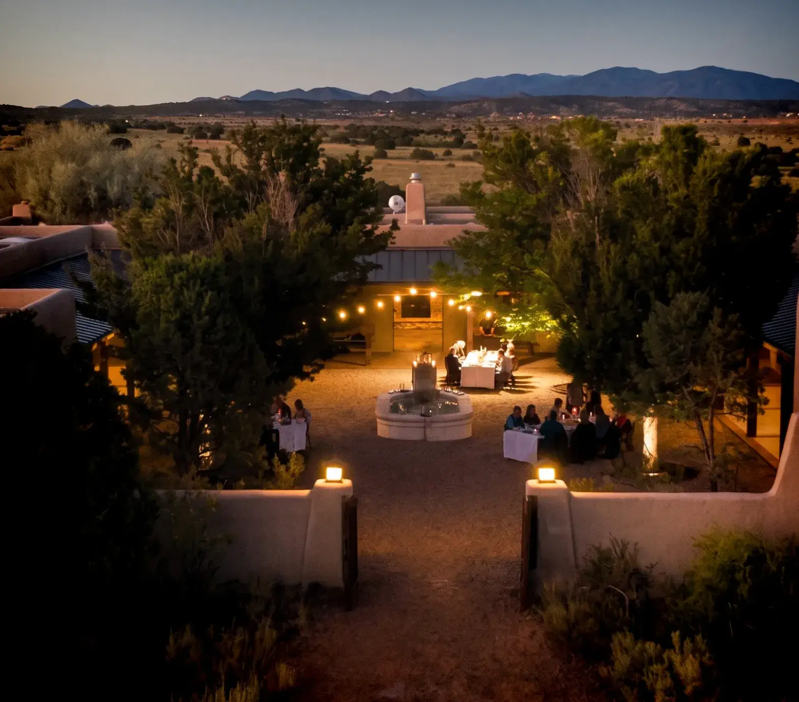 Twilight scene of an outdoor gathering with dining tables, a fountain, and mountains in the background.