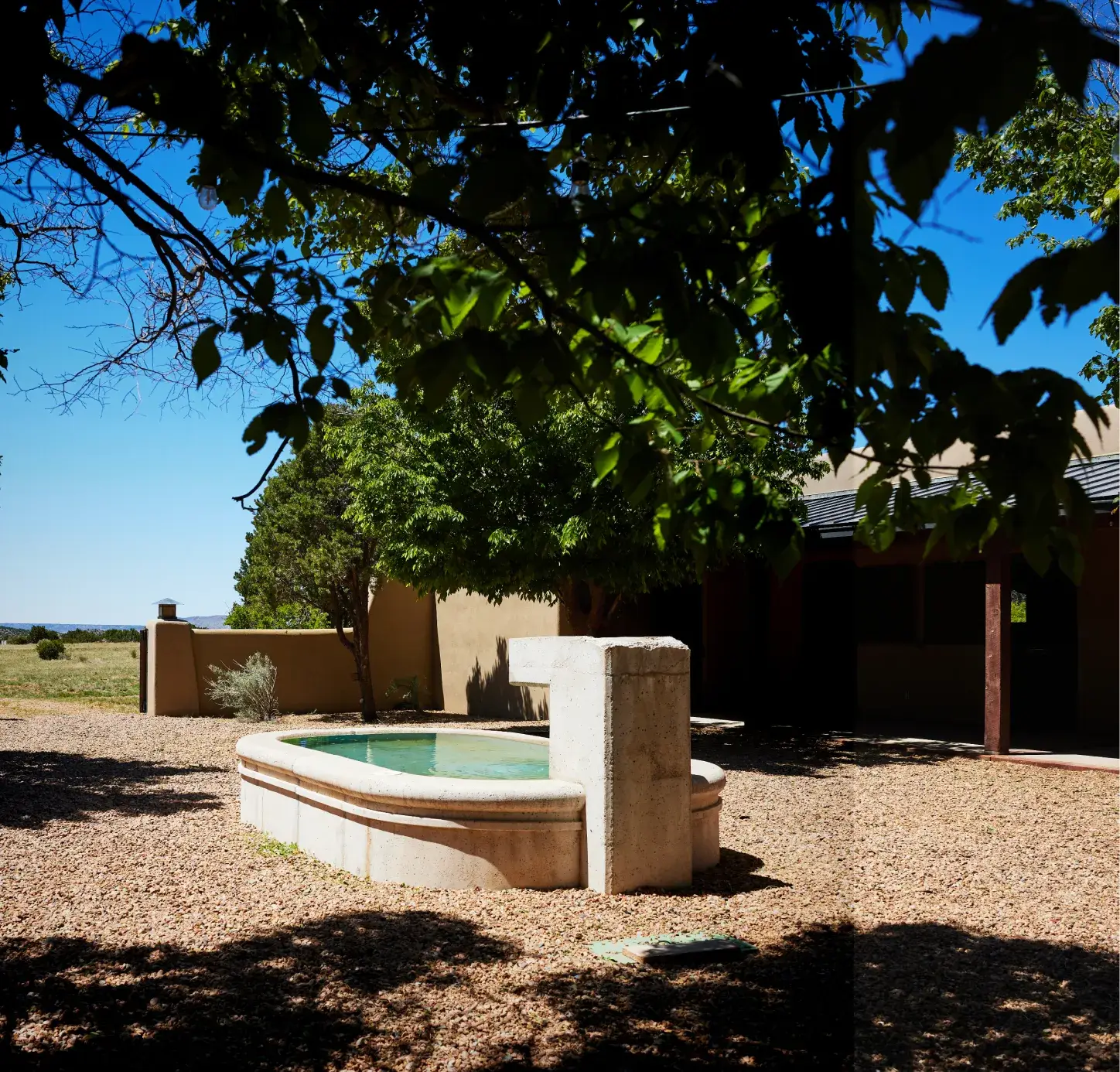 A serene outdoor scene featuring a stone fountain surrounded by trees and gravel, with a clear blue sky in the background.