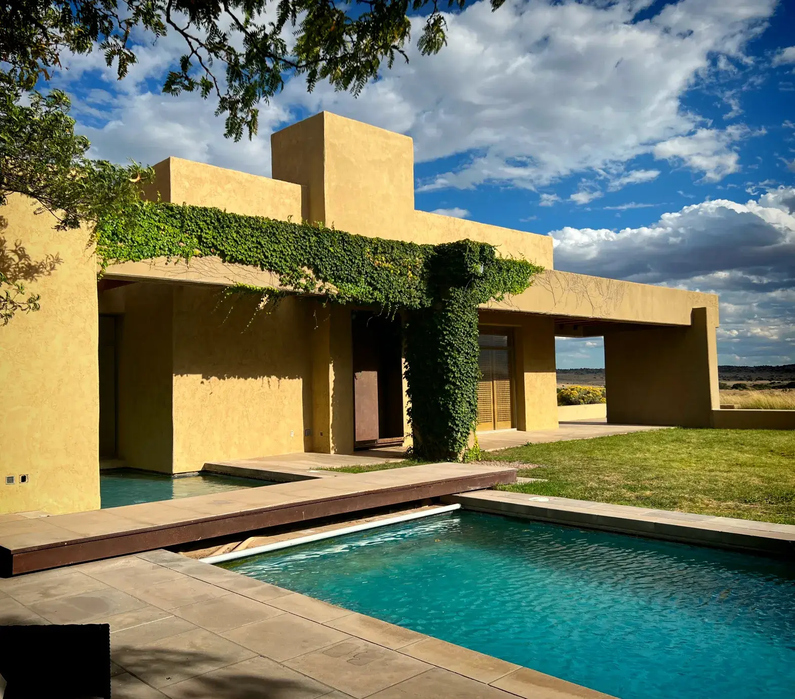 Modern adobe-style house with lush greenery, a swimming pool, and a scenic sky backdrop.