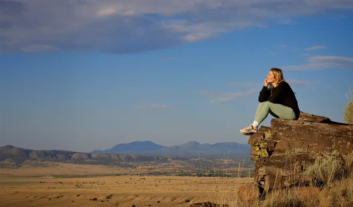 A woman sits thoughtfully on a rock, overlooking a vast landscape with mountains and clear blue skies.