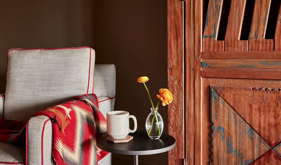 Cozy corner with a gray armchair, colorful blanket, coffee mug, and a vase of flowers beside a rustic wooden door.