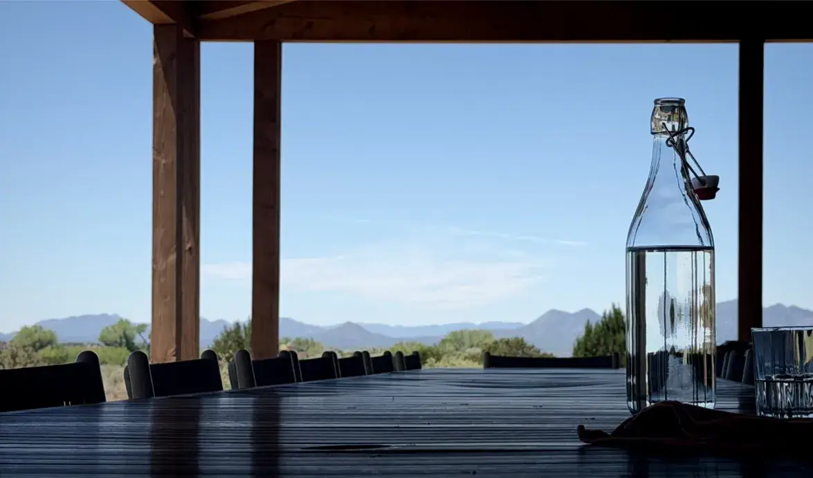 A clear glass bottle of water sits on a wooden table with a scenic mountain view in the background.