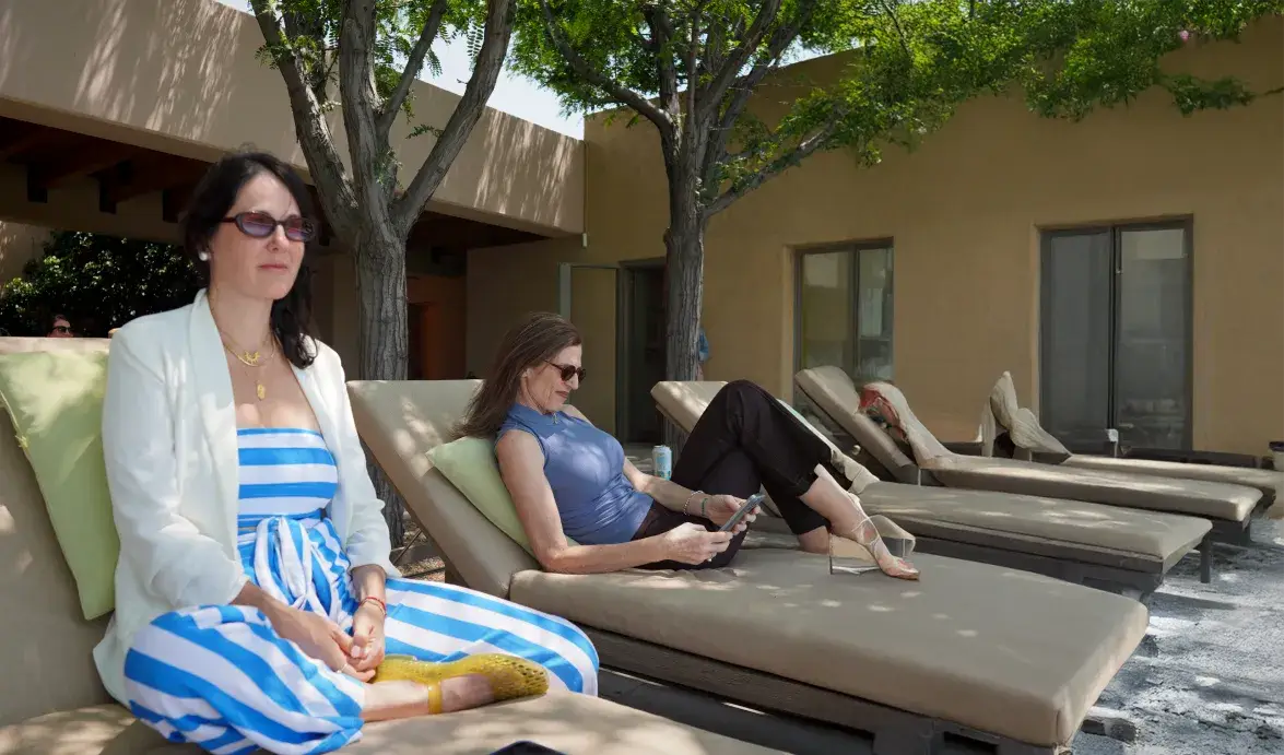 Two women relax on lounge chairs by a pool, one meditating and the other using a tablet in a sunny outdoor setting.