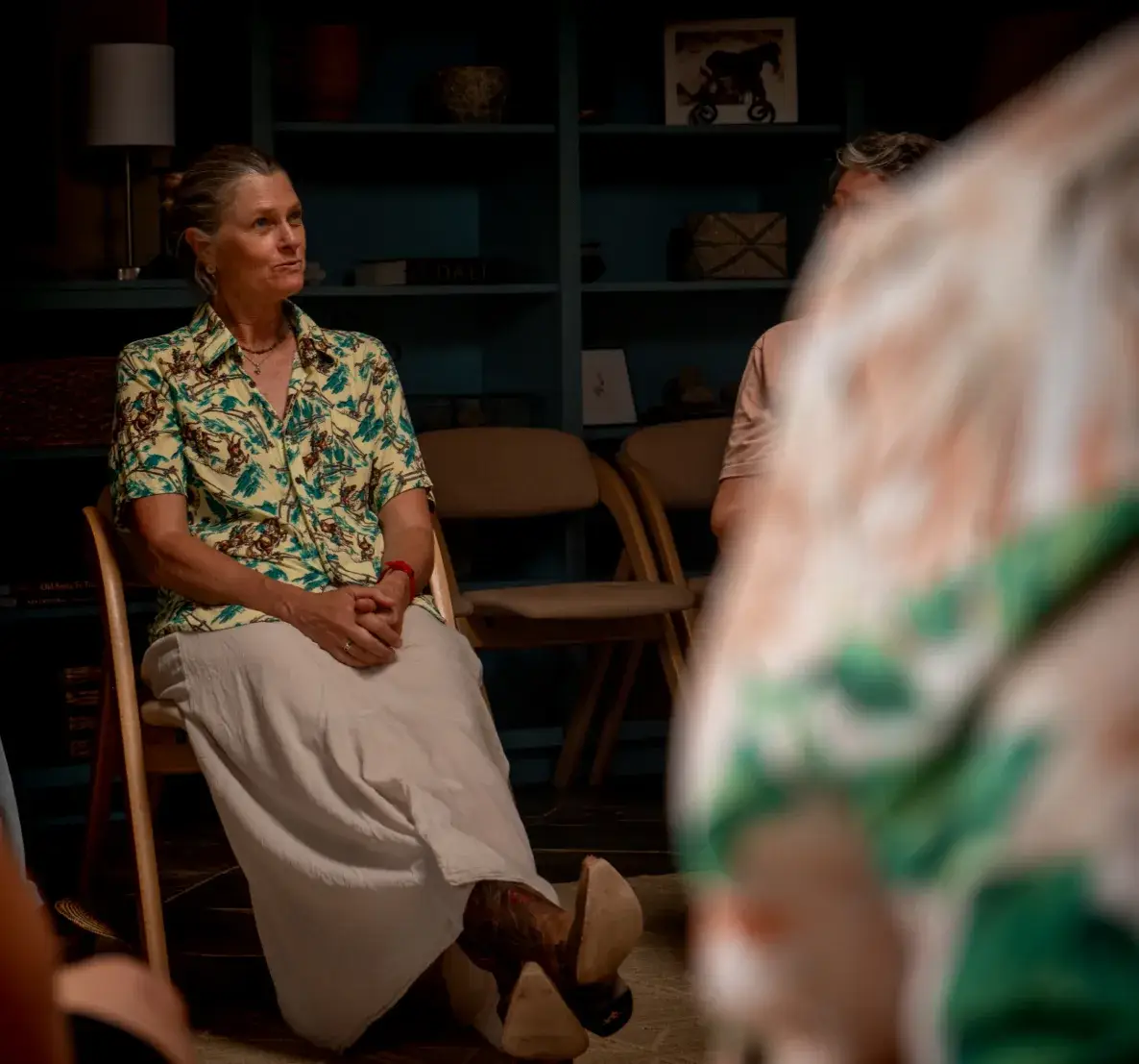 A woman in a colorful shirt sits attentively in a group discussion, surrounded by soft lighting and wooden chairs.