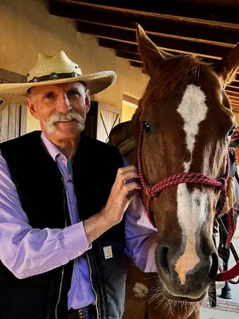 A smiling man in a cowboy hat stands beside a brown horse, gently holding its reins in a rustic stable setting.