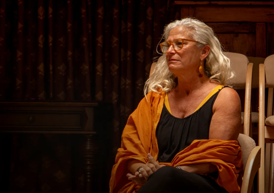 A woman with long gray hair and glasses sits thoughtfully, draped in an orange shawl against a rich, patterned backdrop.