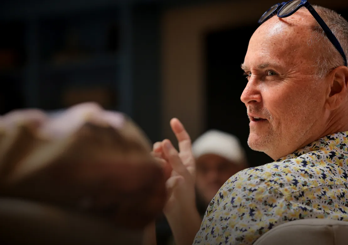 A man in a floral shirt gestures while engaged in conversation with others in a softly lit setting.