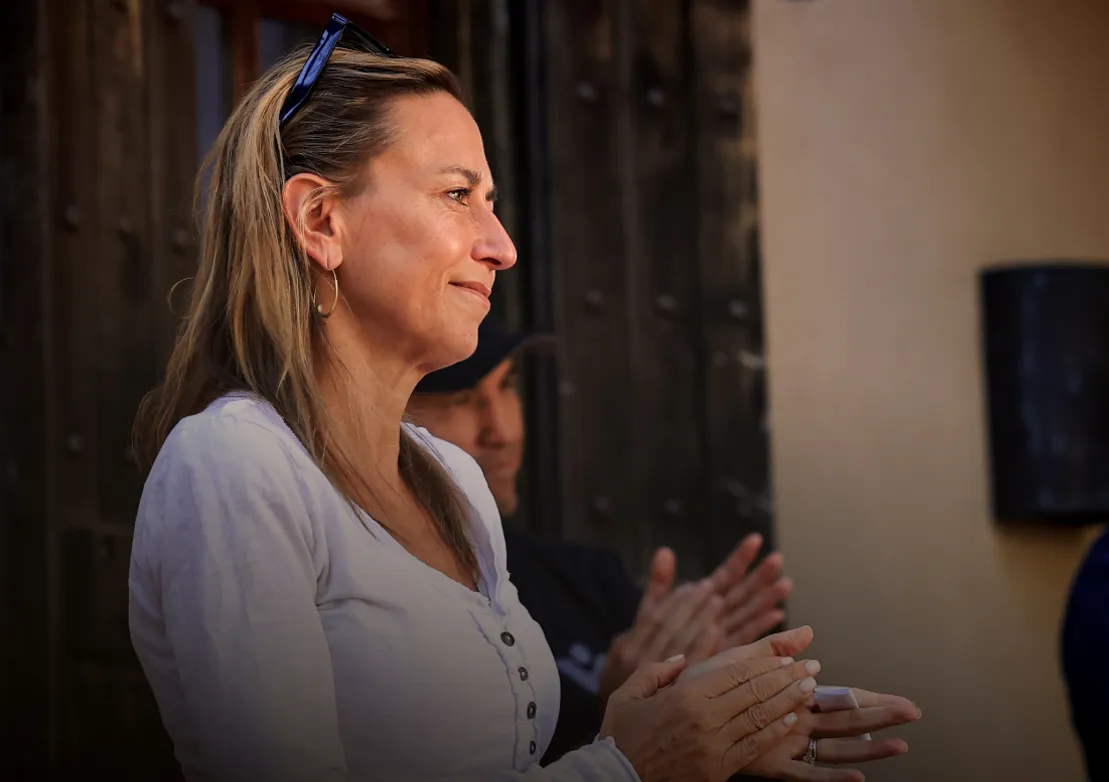 A woman with long hair smiles and claps, showing appreciation during a heartfelt moment at an event.