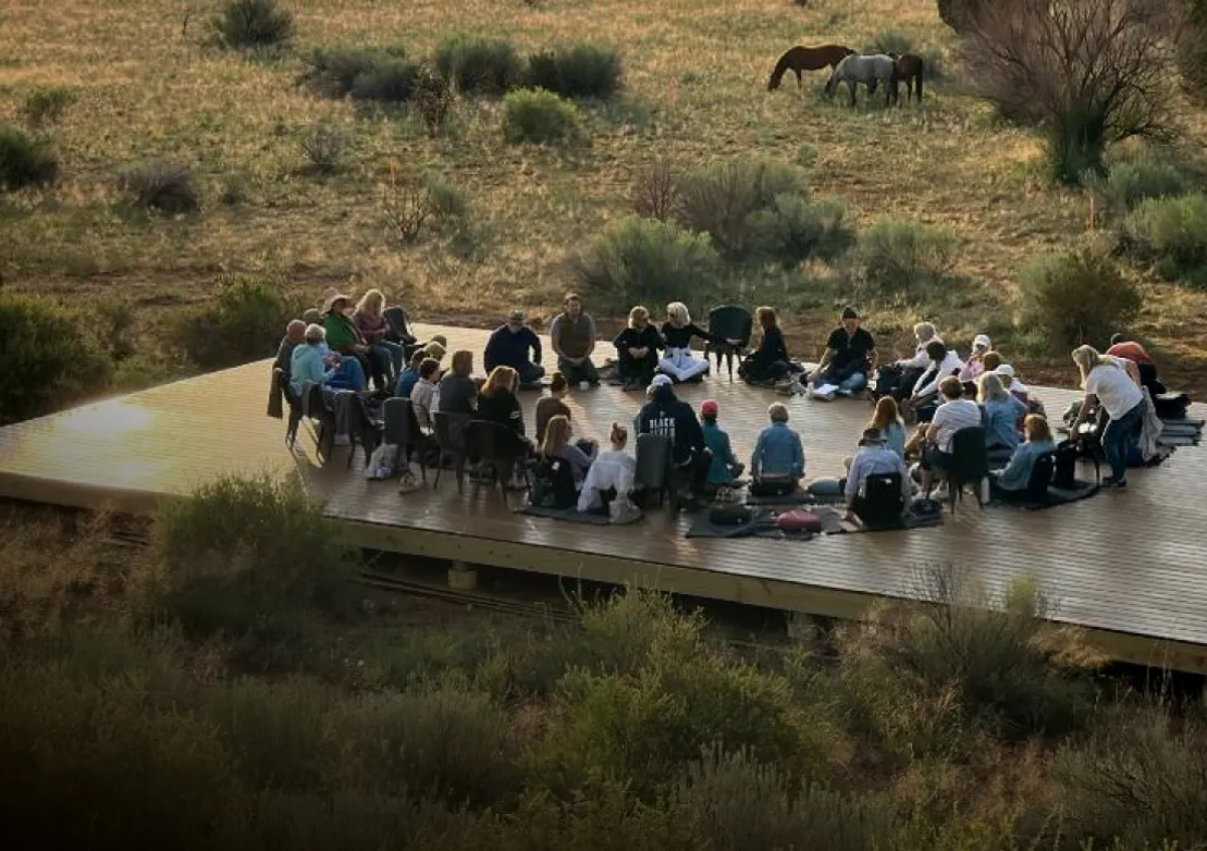 A large group of people gathers on a wooden platform in a natural setting for a workshop or meditation session.