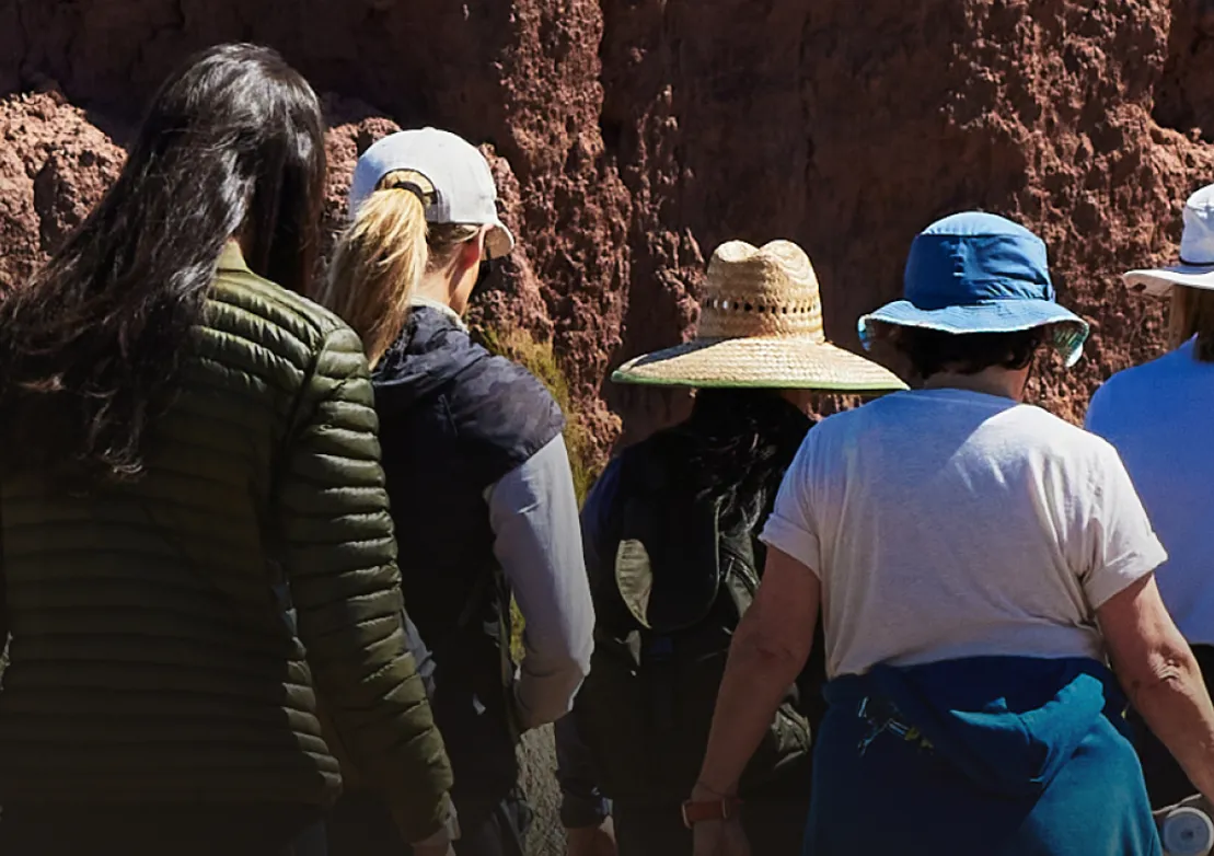 A group of hikers wearing hats and jackets walks along a rocky trail, enjoying a sunny outdoor adventure.