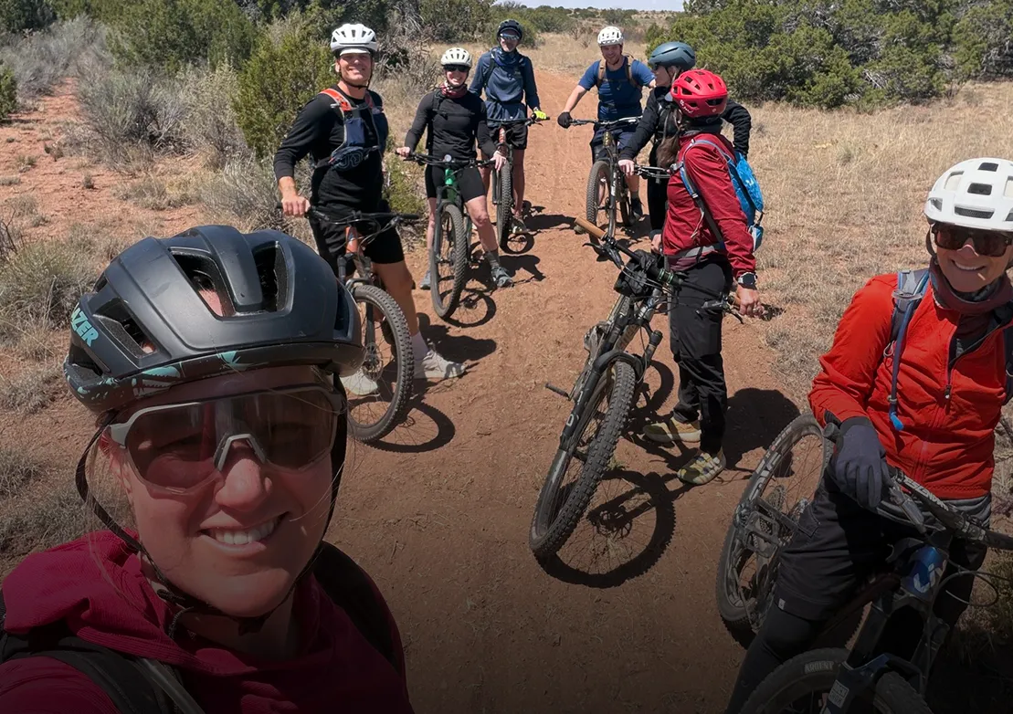 A group of mountain bikers pauses on a dirt trail surrounded by greenery, smiling and enjoying their outdoor adventure.