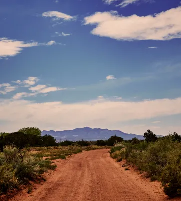 A winding dirt road leads toward distant mountains under a partly cloudy sky, surrounded by desert vegetation.